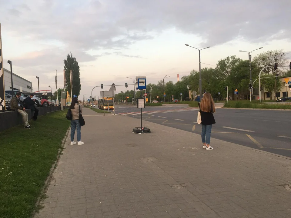 Podróżni MPK Łódź oczekują na autobus na przystanku bez wiaty, ławki i śmietnika.